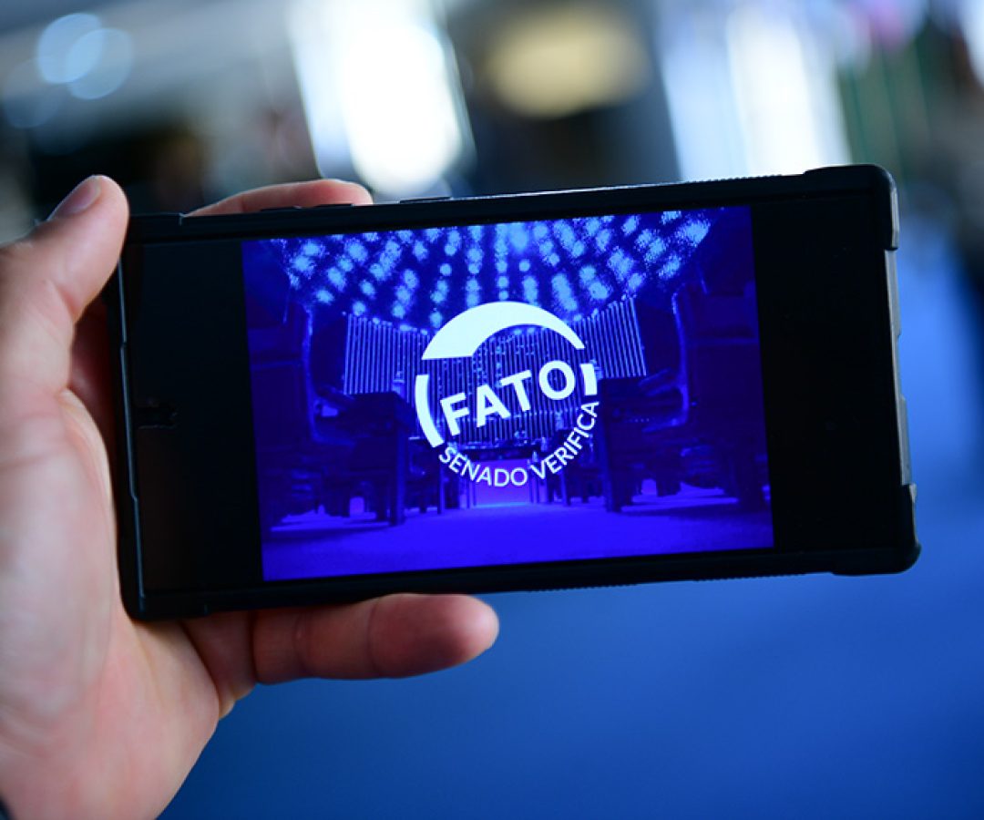Em foco: Uma mão segurando um celular em que a tela mostra um carimbo de Fato.