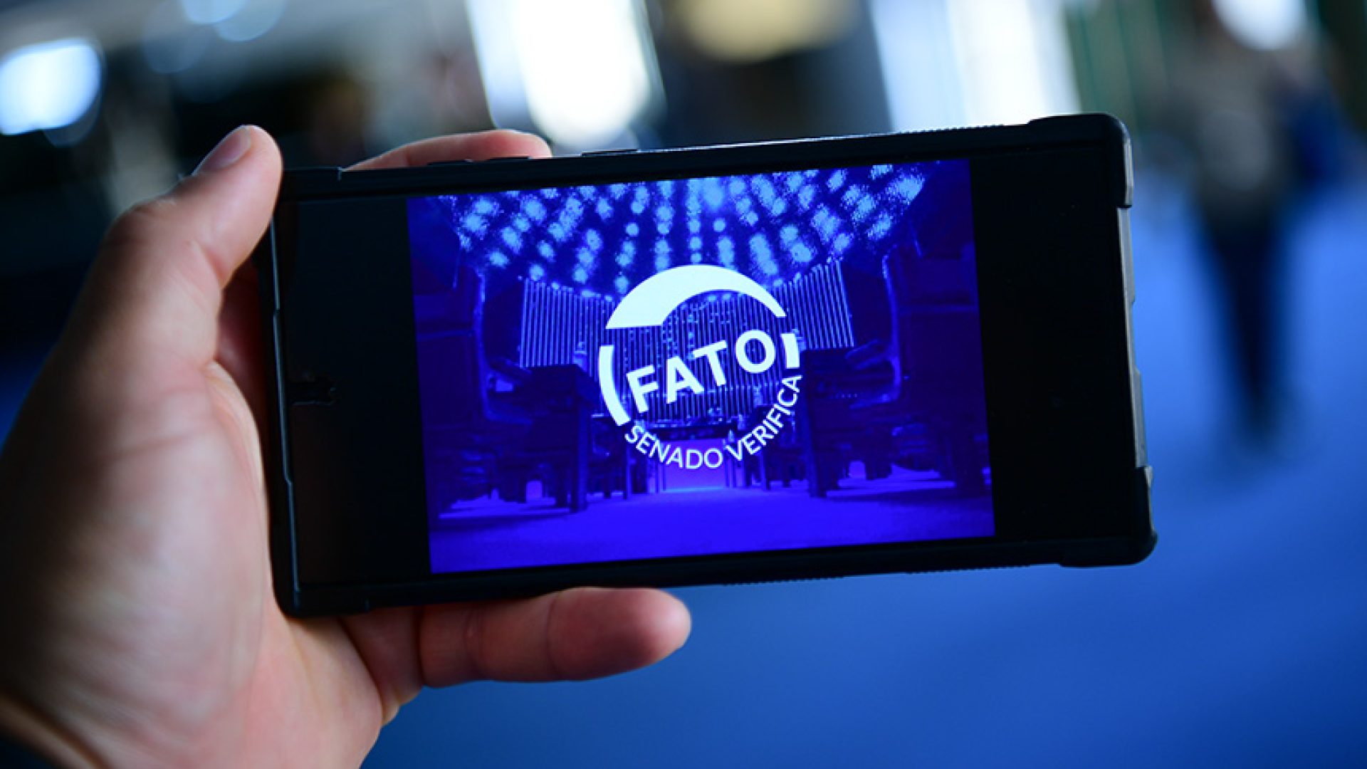 Em foco: Uma mão segurando um celular em que a tela mostra um carimbo de Fato.