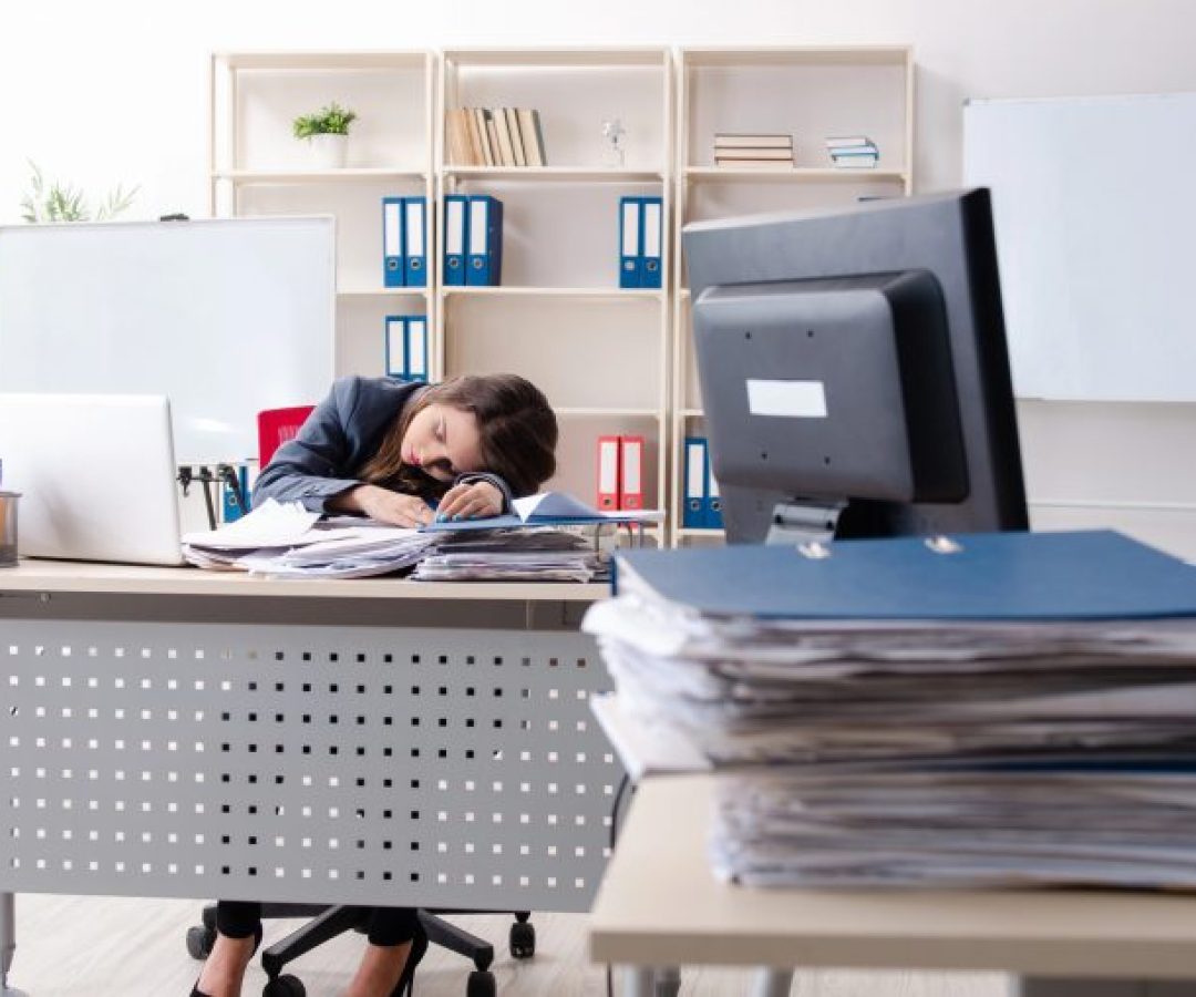 Mulher dorme em com a cabeça em cima da mesa de trabalho, que está cheia de papéis