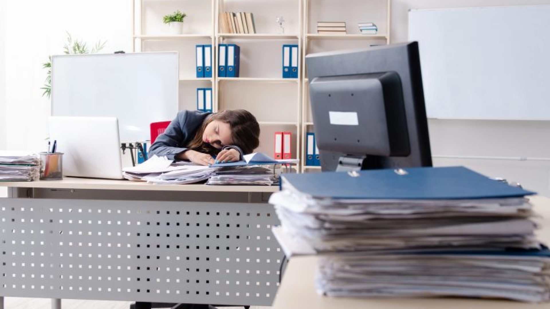 Mulher dorme em com a cabeça em cima da mesa de trabalho, que está cheia de papéis