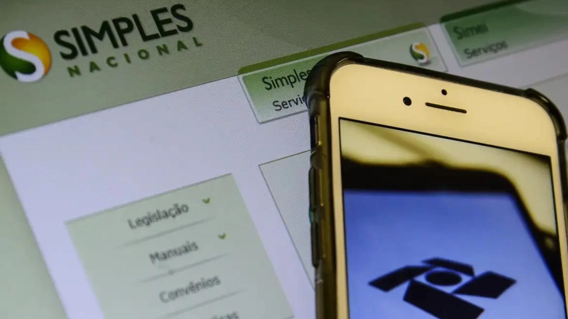 Fotografia de um celular com o aplicativo da receita federal aberto. Ao fundo, vemos, em uma tela de computador, o site do Simples Nacional