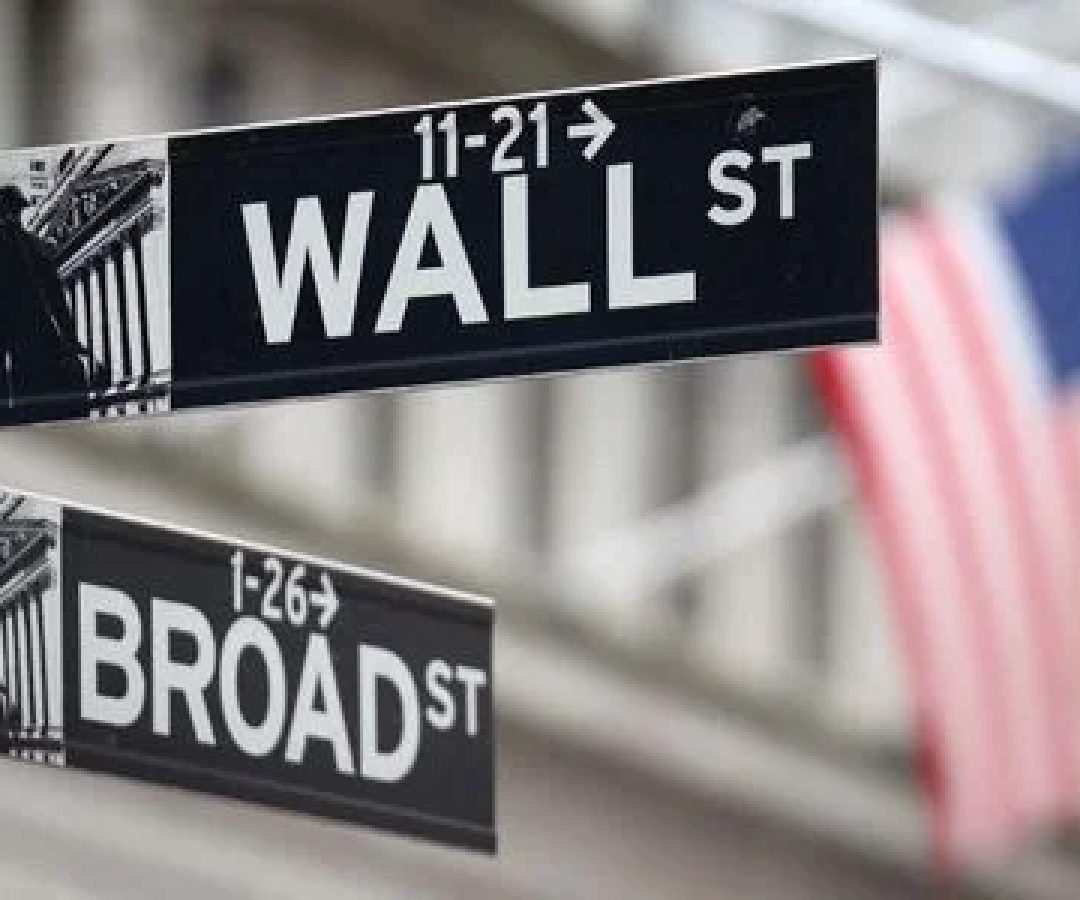 Placa de Wall Street em frente a bandeira dos EUA, do lado de fora da Bolsa de Valores de Nova York; investir