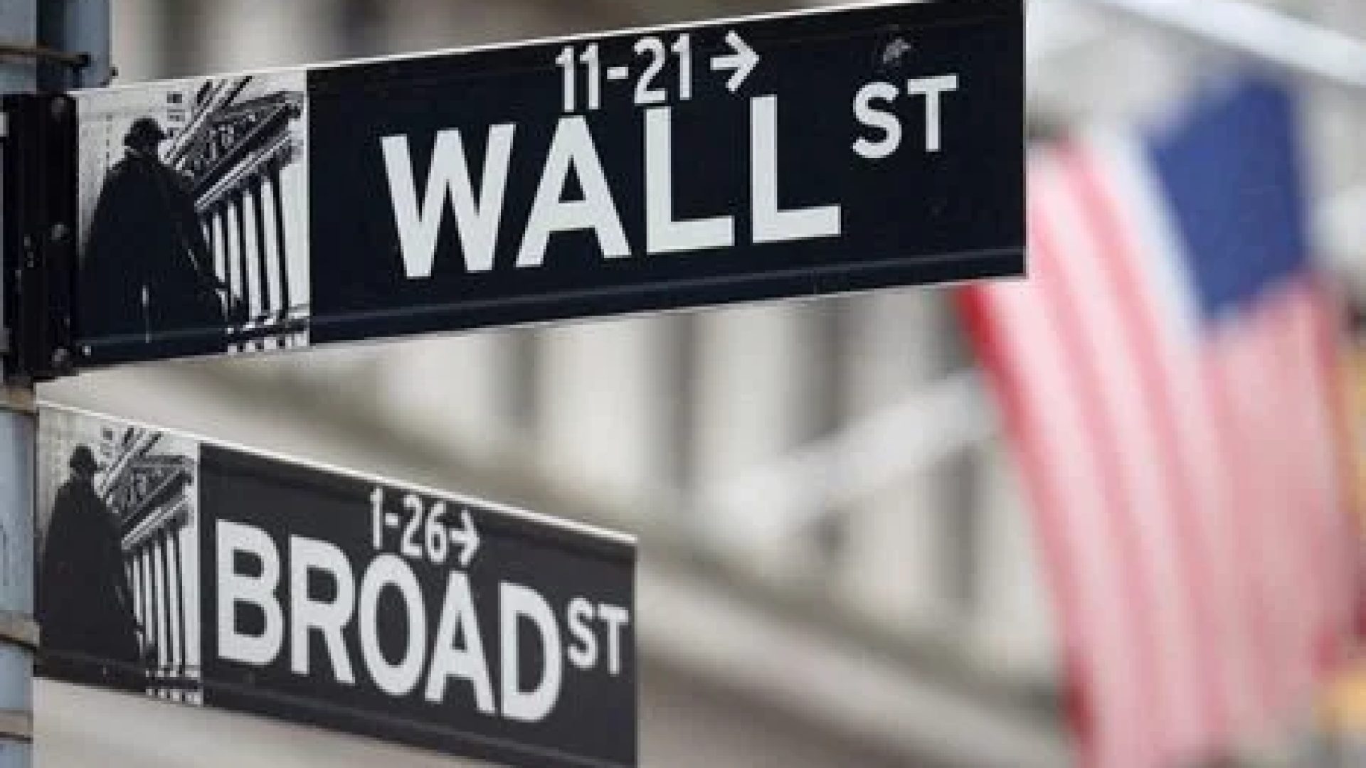 Placa de Wall Street em frente a bandeira dos EUA, do lado de fora da Bolsa de Valores de Nova York; investir